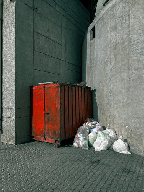 A black cylindrical bin with a partially open white plastic lid, situated against a large, smooth, beige-toned stone wall in an indoor or outdoor urban environment. The bin is overflowing with various