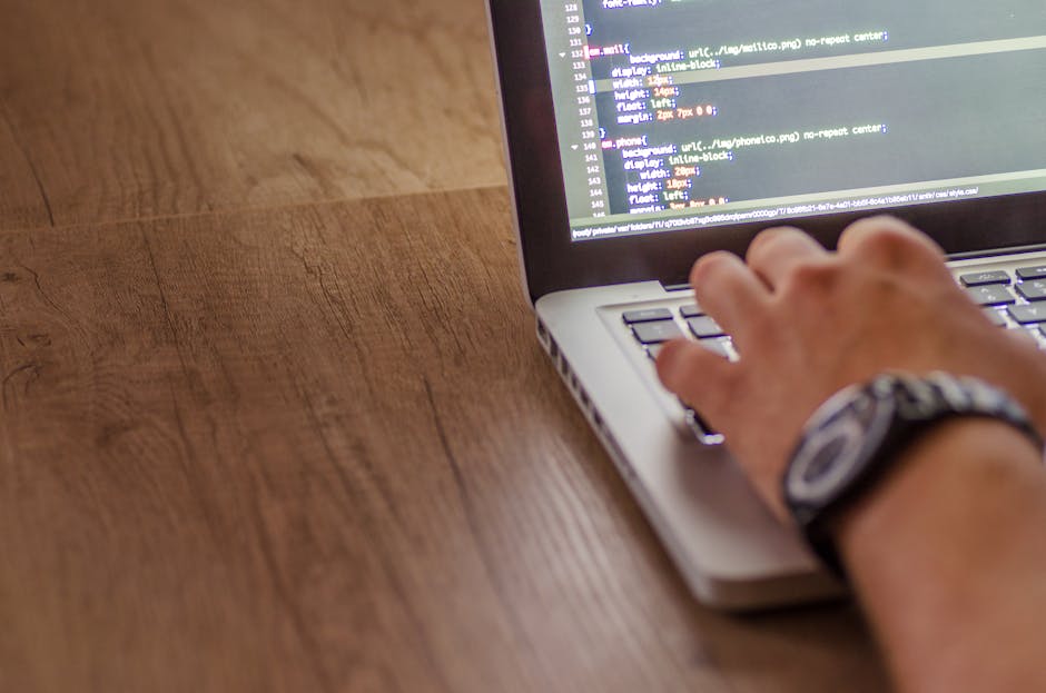 A person with a metallic wristwatch typed on a silver laptop placed on a wooden table surface. The laptop screen displays lines of code with a dark background and multicolored text, indicating program